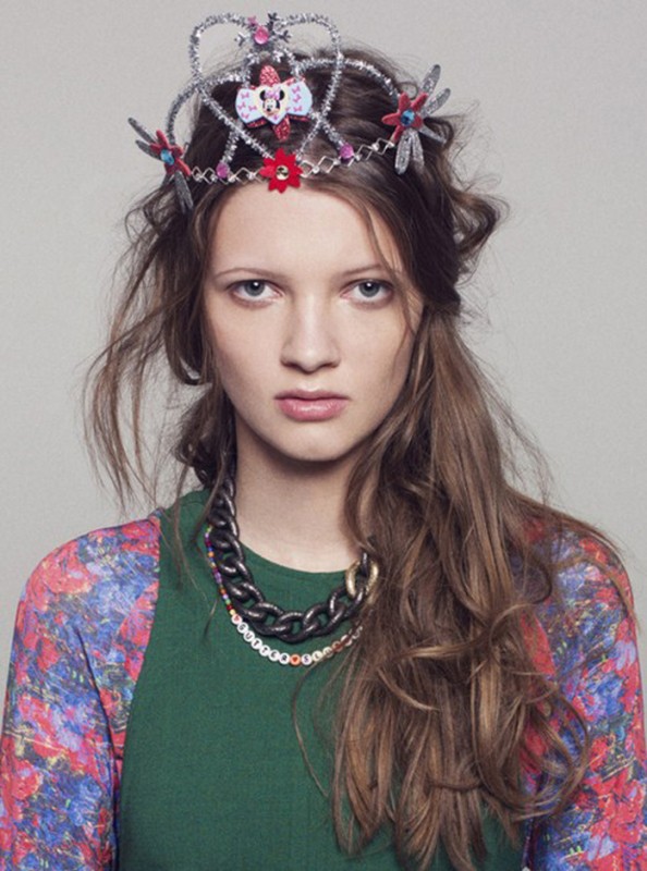 Portrait of a young woman wearing a decorative crown and chunky necklace, styled with a floral-patterned top, against a light background.