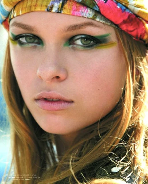 Close-up shot of a woman with fair skin, wearing colorful eye makeup and a patterned turban-style head covering; beauty advertisement.