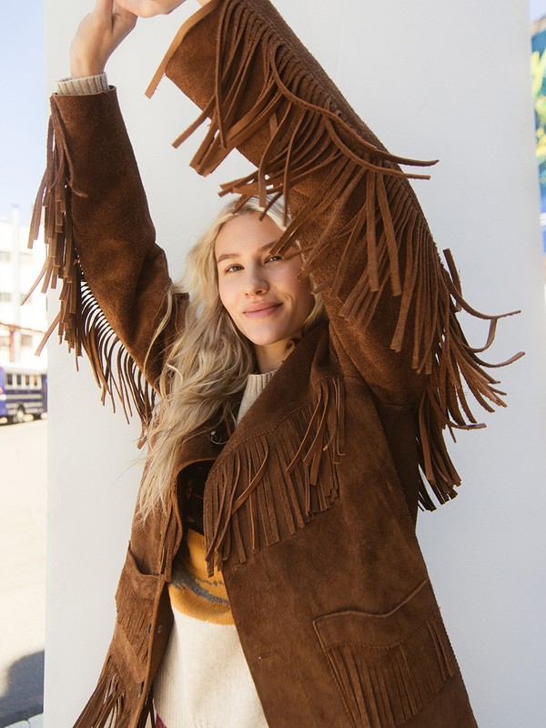 Adult female posing outdoors in a brown suede jacket with fringes, cream sweater, with blonde hair and neutral facial expression.