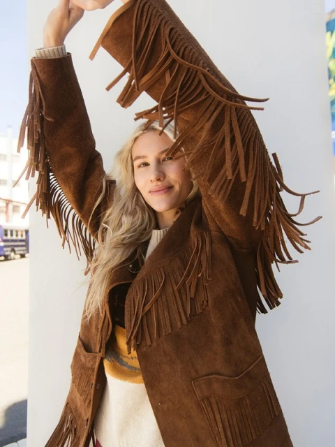 Adult female posing outdoors in a brown suede jacket with fringes, cream sweater, with blonde hair and neutral facial expression.
