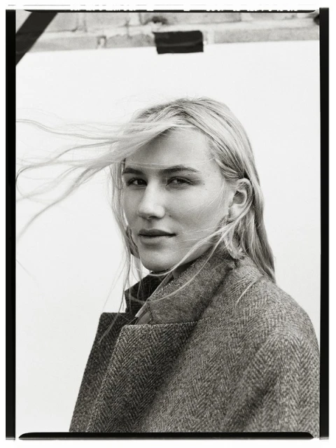 Portrait of a blonde woman in a herringbone coat with windswept hair. She poses in front of a neutral background with partial brick.