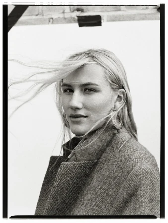 Portrait of a blonde woman in a herringbone coat with windswept hair. She poses in front of a neutral background with partial brick.