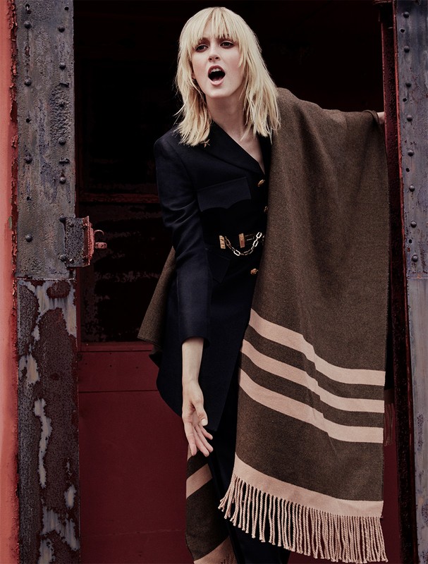 Blonde woman wearing a black jacket and striped brown blanket shawl standing in a dark doorway