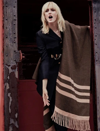 Blonde woman wearing a black jacket and striped brown blanket shawl standing in a dark doorway