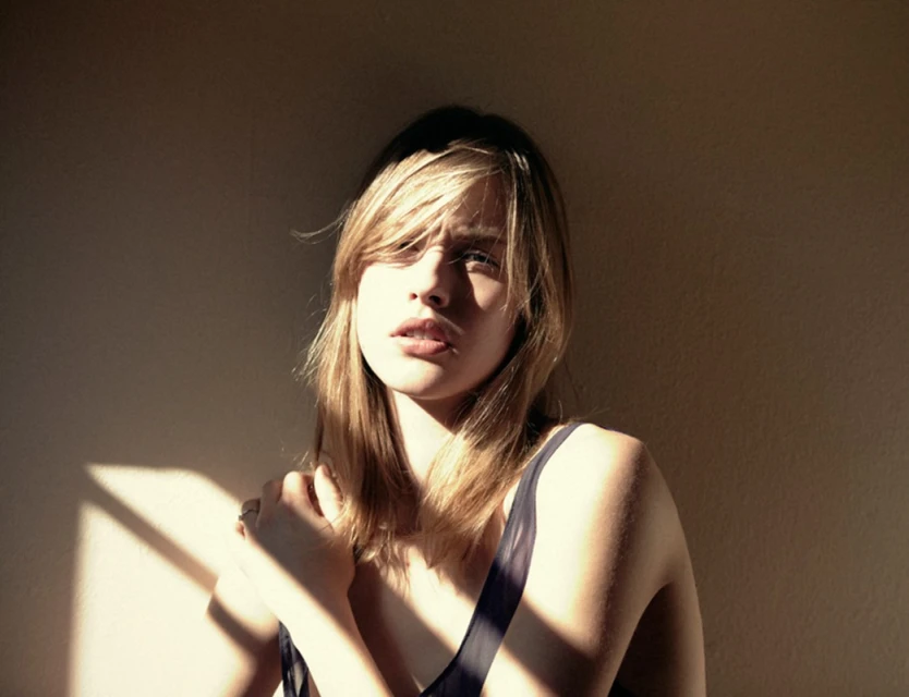 Portrait of a young adult woman with long blonde hair in natural light, wearing a dark top, posing indoors with soft shadows on the wall.