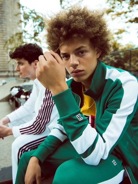 Two young adult males sit outside. One wears a green tracksuit, and the other a white tracksuit with red stripes on the sleeves.