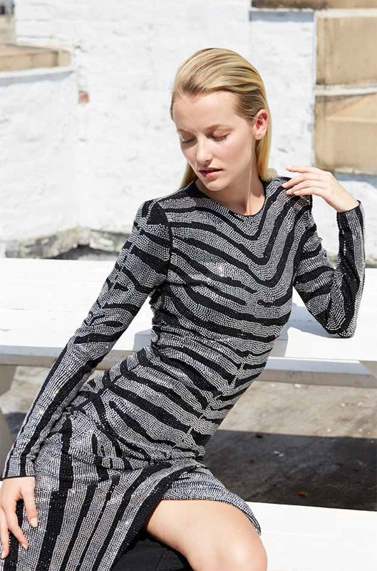 A blonde adult woman in a silver and black patterned dress poses against a light brick background outdoors, looking down with a calm facial expression.