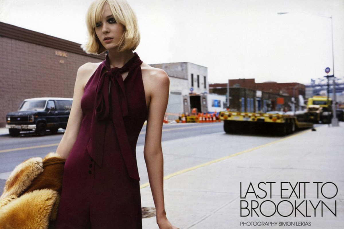 A stylish blonde woman in a burgundy jumpsuit stands on a city street, holding a fur coat with buildings, truck, and van in background.
