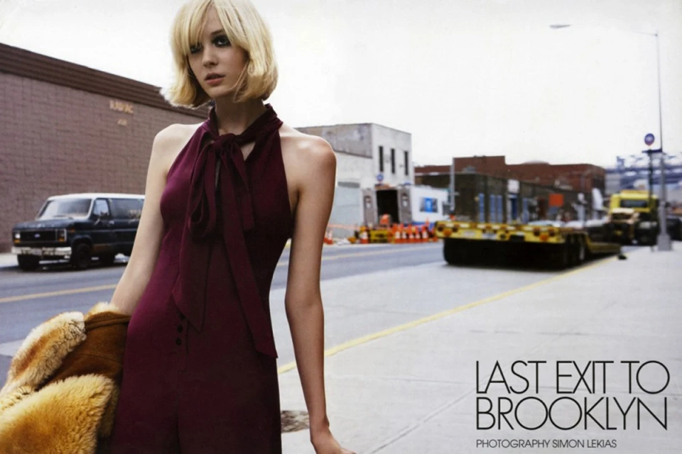 A stylish blonde woman in a burgundy jumpsuit stands on a city street, holding a fur coat with buildings, truck, and van in background.