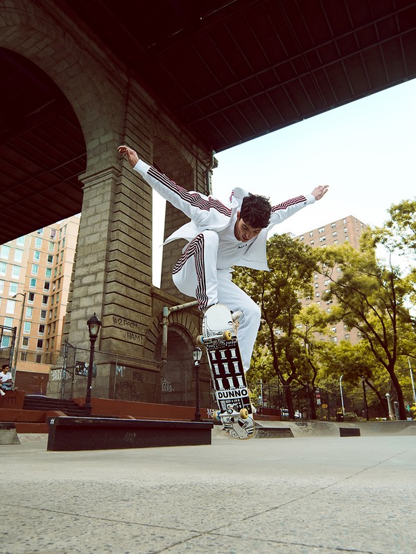 Man in white suit mid-air on skateboard under bridge, performing a stunt; cityscape and blue sky in background, fashion, sport, lifestyle.