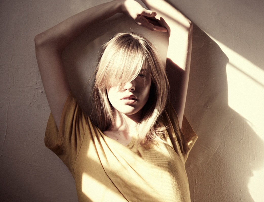 Woman in a yellow t-shirt with her arms raised overhead standing against a textured wall with hard directional window light