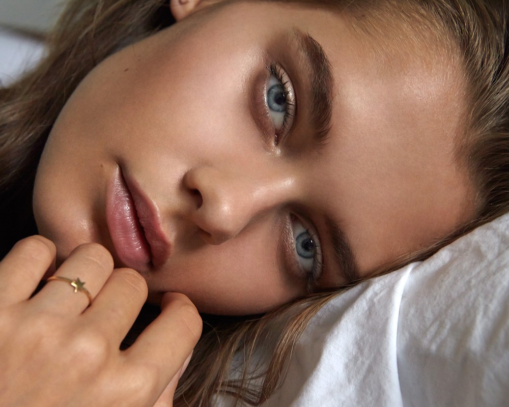 Close-up of a fair-skinned young adult woman with blue eyes, lying on a white pillow, wearing a gold star ring, beauty shot.