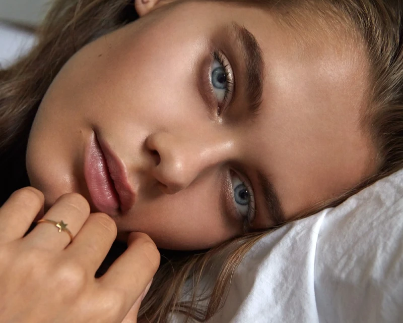 Close-up of a fair-skinned young adult woman with blue eyes, lying on a white pillow, wearing a gold star ring, beauty shot.
