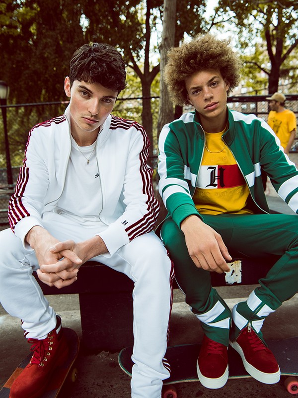 Two young men sit on a skateboard while modeling tracksuits in an urban park setting with green trees and bright daylight.