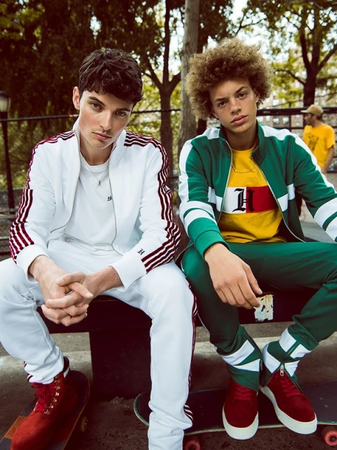 Two young men sit on a skateboard while modeling tracksuits in an urban park setting with green trees and bright daylight.