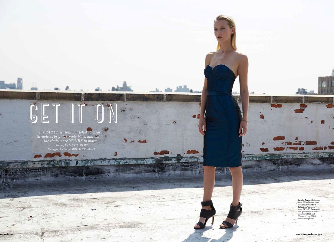 Blonde fashion model poses in a strapless blue dress and black high heels against an urban rooftop and city background in a fashion editorial.