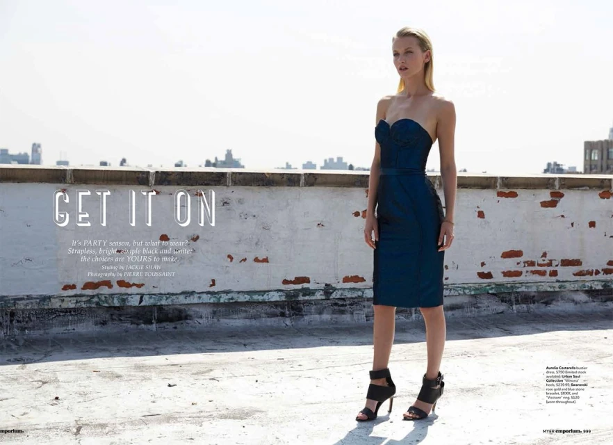 Blonde fashion model poses in a strapless blue dress and black high heels against an urban rooftop and city background in a fashion editorial.