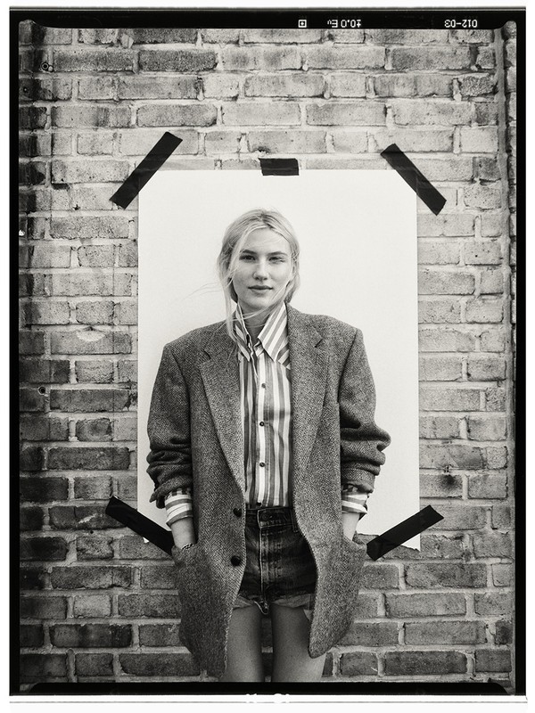 Monochrome portrait of a woman in a striped shirt and oversized blazer, posing against a brick wall with a white paper backdrop.