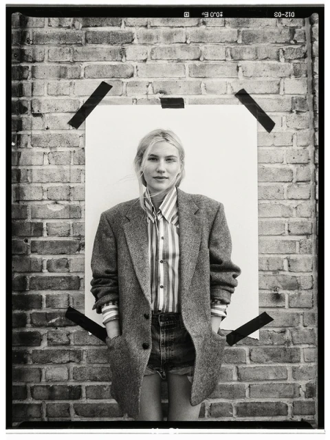 Monochrome portrait of a woman in a striped shirt and oversized blazer, posing against a brick wall with a white paper backdrop.