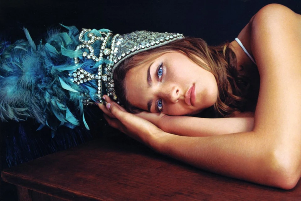 Woman with feather headpiece rests head on a wood surface in a dimly lit setting, portrait orientation. Glamorous fashion headwear.