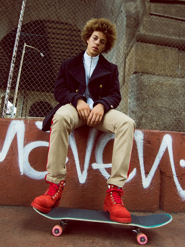 Young man with curly hair in a dark coat and khaki pants sits on a skateboard against a graffiti wall outdoors in an urban setting.