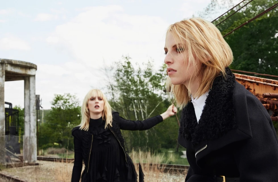 Two adult women pose outdoors wearing fashionable black coats on a cloudy day. One woman is partially obscured in the background.