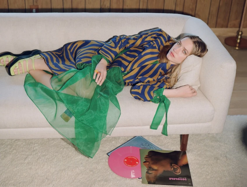 A blonde woman in a patterned dress reclines on a cream couch next to vinyl records in a brightly lit indoor setting.
