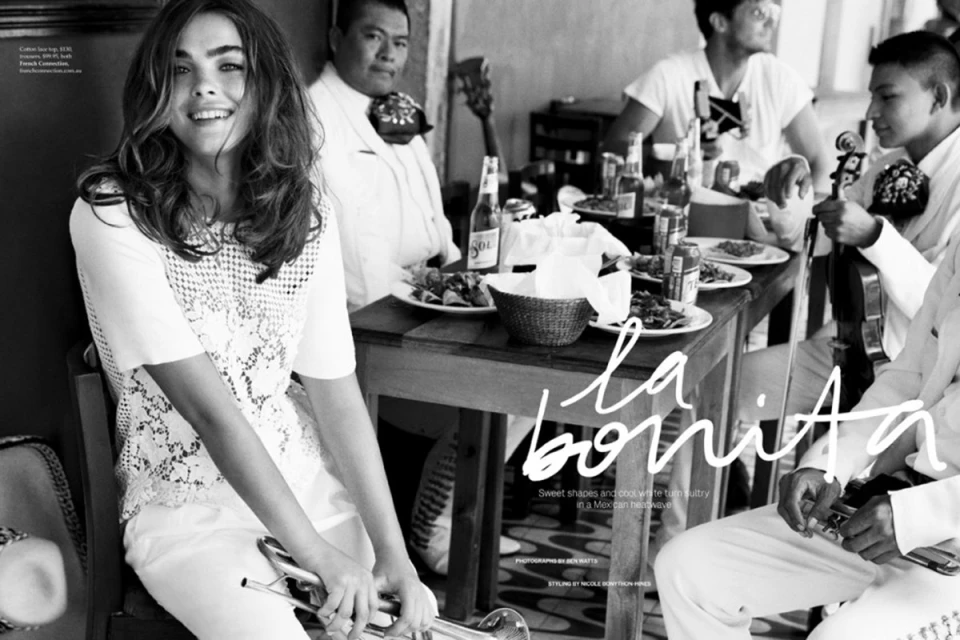 Black and white image featuring a smiling woman in a lace top, seated with a trumpet, surrounded by musicians at a table.