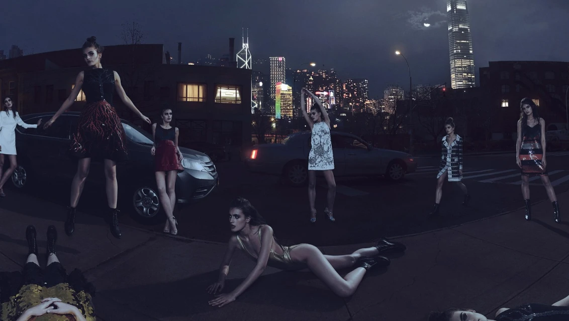 Fashion photo shoot featuring multiple adult female models in short dresses, posing outside at night with cars and a city skyline in the background.