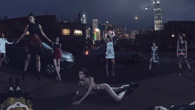 Fashion photo shoot featuring multiple adult female models in short dresses, posing outside at night with cars and a city skyline in the background.