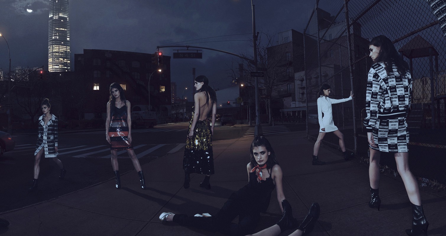Fashion editorial shoot at night with six adult female models posing in streetwear on a city street with urban buildings in the background.