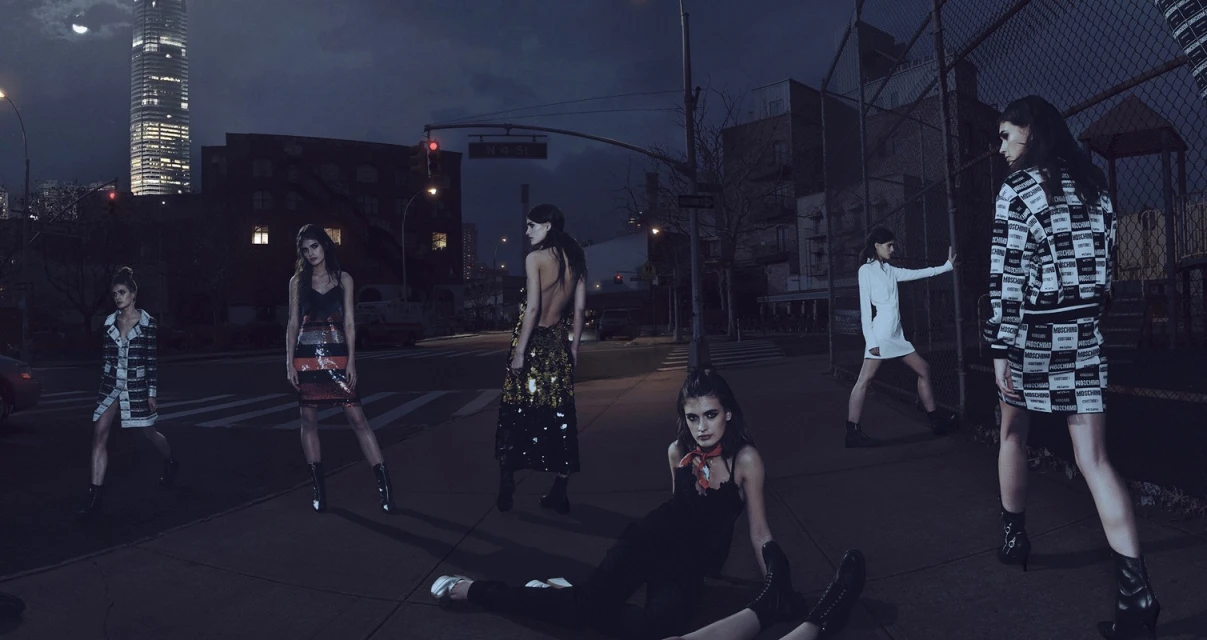 Fashion editorial shoot at night with six adult female models posing in streetwear on a city street with urban buildings in the background.