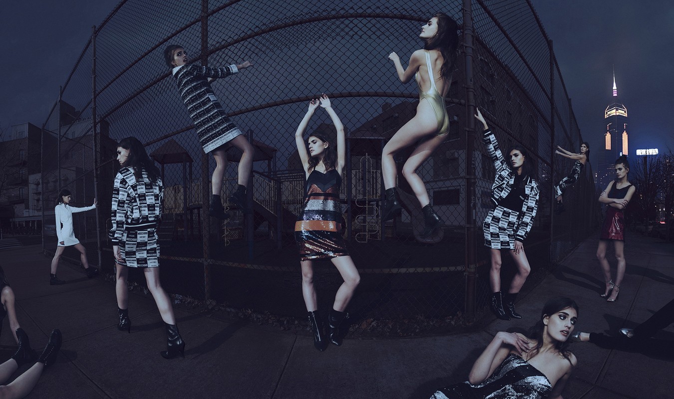 Fashion photoshoot featuring several women in various poses and outfits against a chain-link fence and blurred city skyline at night.