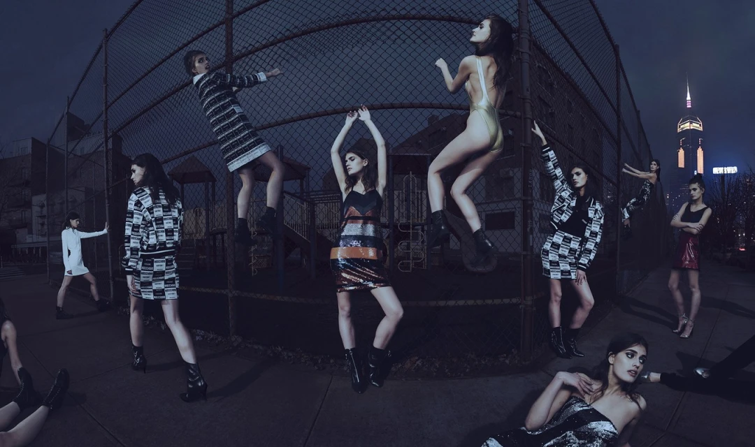 Fashion photoshoot featuring several women in various poses and outfits against a chain-link fence and blurred city skyline at night.