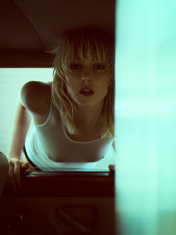 Woman with blonde hair and a white tank top leans forward indoors, with a diffused light creating a soft, ethereal look.