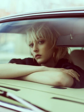 Blonde woman resting her arms on a car dashboard, viewed through the windshield.