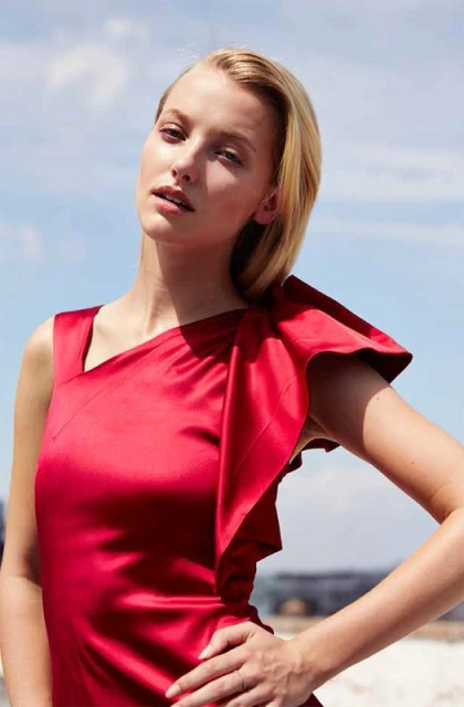 A beautiful fair-skinned blonde woman in a red satin one-shoulder dress poses outdoors with blue sky and clouds in the background.
