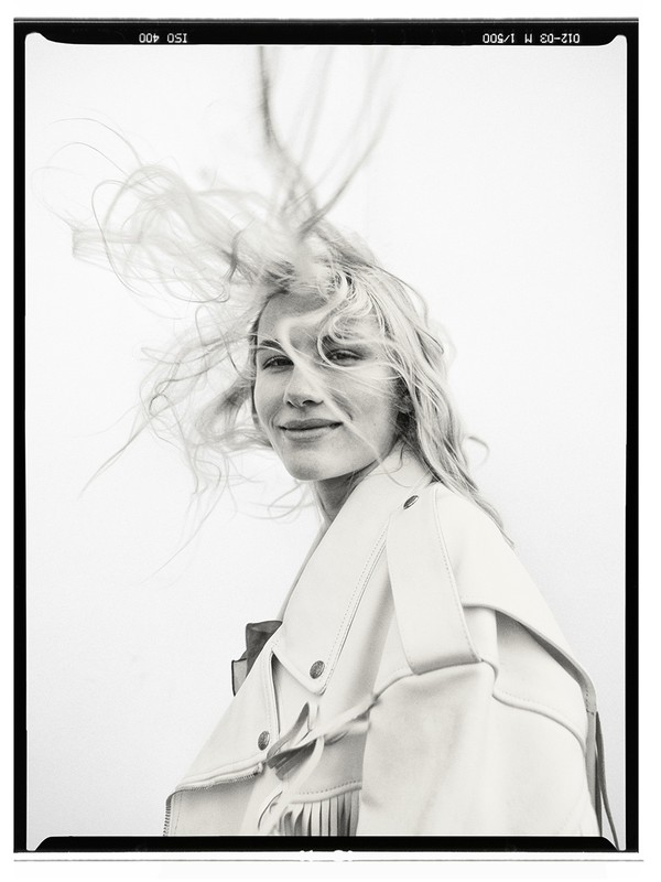 Portrait of a blonde woman with hair blowing in the wind wearing a fringed jacket against a light background