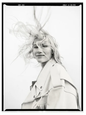 Portrait of a blonde woman with hair blowing in the wind wearing a fringed jacket against a light background