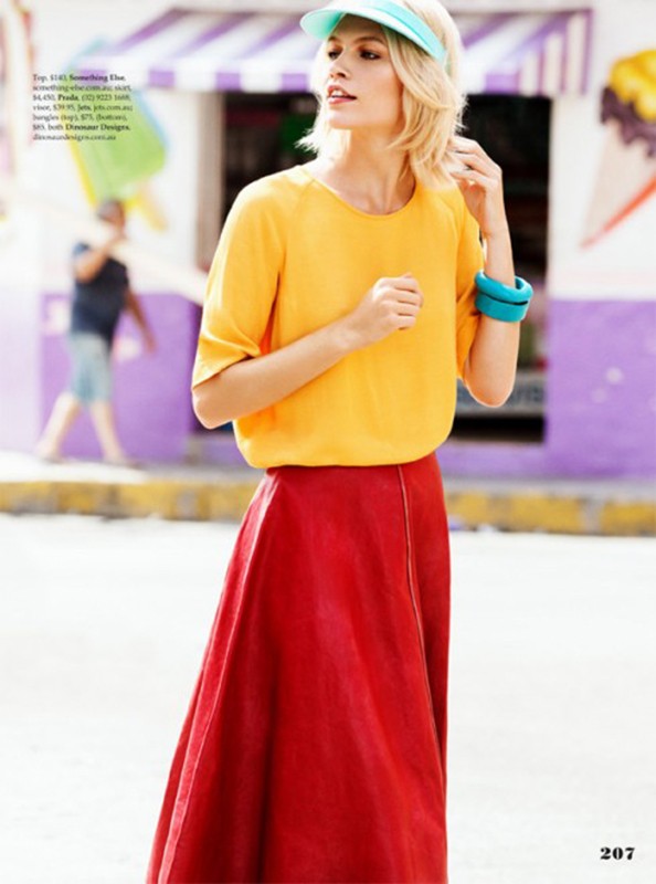 Blonde woman wearing a yellow blouse, red skirt, and turquoise visor outdoors, with a person walking in the background.
