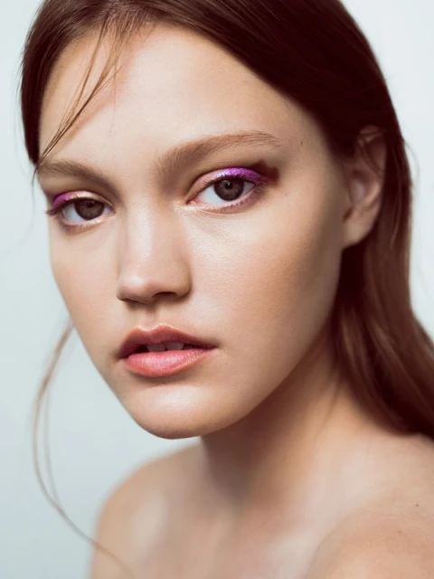 Close-up portrait of a young adult female model with auburn hair, pink eye makeup, fair skin, and light pink lips against a white background.