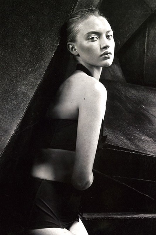 Black and white portrait of a woman in a two-piece outfit, looking at the camera, leaning against a dark, geometric background.