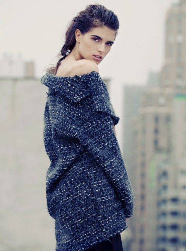 Woman in tweed jacket poses against a blurry cityscape. The jacket has an off-the-shoulder design and textured look, with a neutral backdrop.