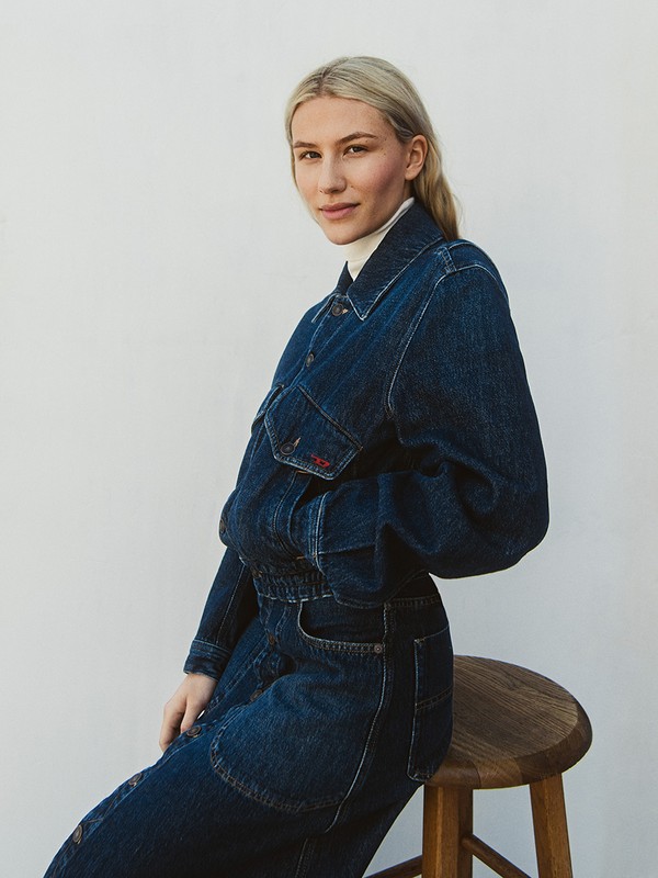 Blonde woman wearing a dark blue denim jacket and jeans seated on a wooden stool against a white wall.