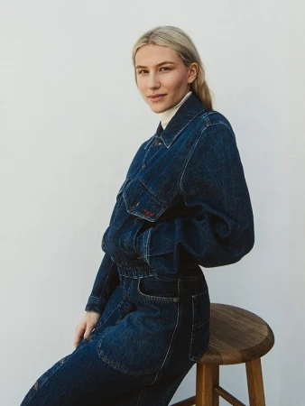 Blonde woman wearing a dark blue denim jacket and jeans seated on a wooden stool against a white wall.