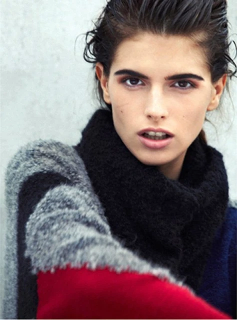 Headshot of a woman with dark hair and fair skin wearing a colorful knit sweater against a neutral gray background in soft lighting.