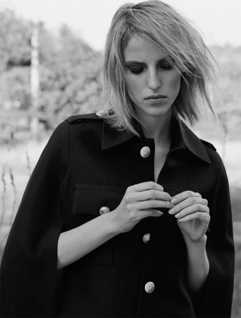 Black and white portrait of an adult woman wearing a dark jacket with metal buttons outdoors, looking down and adjusting her jacket.
