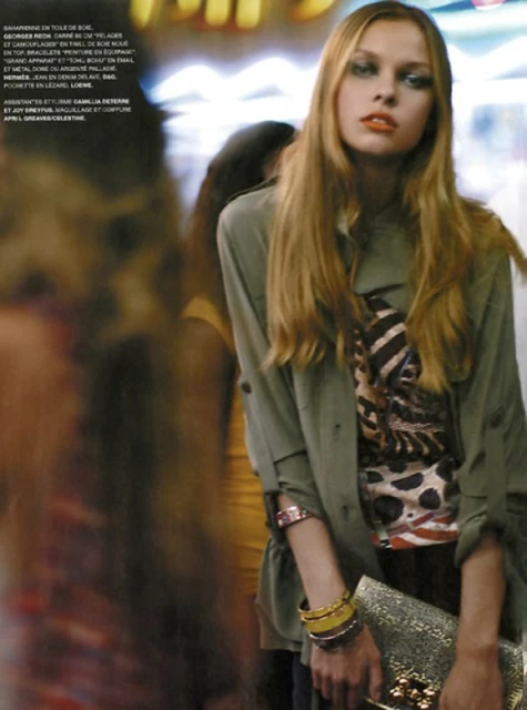 Fashion editorial shot of a fair-skinned blonde model in an olive jacket, animal-print top, holding a gold clutch, posing indoors with blurred figures.