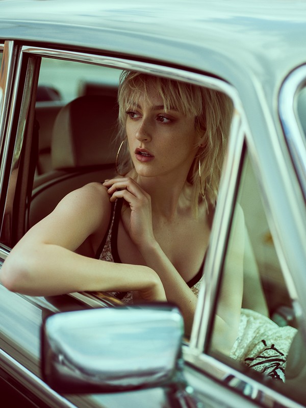 Stylish blonde woman inside a vintage car, glancing away from the camera. She wears a patterned top and hoop earrings, vintage fashion.