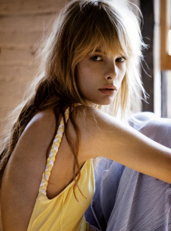 Young woman with blonde hair poses indoors wearing a yellow tank top with braided straps, looking at the camera with neutral expression.